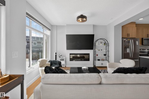 The open concept living area features large windows, hardwood flooring, a modern electric fireplace, and a sleek ceiling light fixture - 28 6214 Cartmell Crest, Edmonton, AB - Indoor Photo Showing Living Room With Fireplace