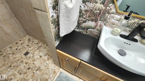 Tiled shower with mosaic floor and large format wall tiles, vanity with wood-finish cabinetry, dark countertop, and an integrated white sink with a black faucet, framed mirror, and decorative wallpaper - 3531 46 Street, Edmonton, AB - Indoor Photo Showing Bathroom
