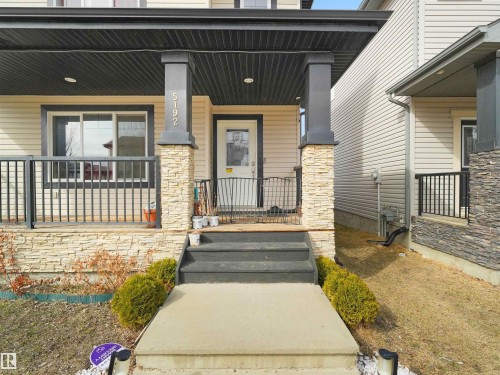 Welcoming front entrance featuring stone-clad pillars, a covered porch with black railings, and a contrasting light-colored siding exterior - 5192 1B Avenue Sw, Edmonton, AB - Outdoor With Deck Patio Veranda