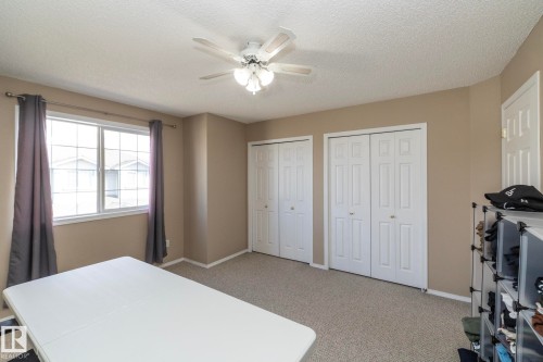 Carpeted room featuring a white ceiling fan with integrated lighting, a multi-pane window, two white bi-fold closet doors, and neutral wall paint - 50 14803 Miller Boulevard, Edmonton, AB - Indoor Photo Showing Bedroom