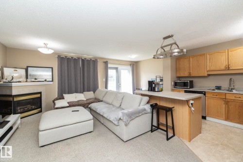 Open-concept living space featuring a corner fireplace with a stone mantel, light-colored carpeting, a kitchen island with seating, natural wood cabinetry, and stainless steel appliances - 50 14803 Miller Boulevard, Edmonton, AB - Indoor With Fireplace