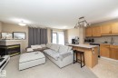 Open-concept living space featuring a corner fireplace with a stone mantel, light-colored carpeting, a kitchen island with seating, natural wood cabinetry, and stainless steel appliances - 50 14803 Miller Boulevard, Edmonton, AB  - Indoor With Fireplace 