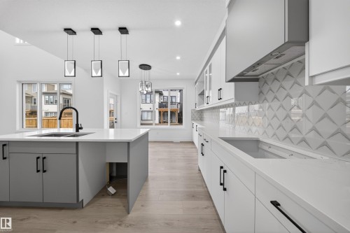 Modern kitchen featuring a large island with an undermount sink and matte black faucet, light wood-finish flooring, white perimeter cabinetry with dark pulls, quartz countertops, and a striking geometric tile backsplash - 3906 41 Avenue, Beaumont, AB - Indoor Photo Showing Kitchen With Double Sink With Upgraded Kitchen