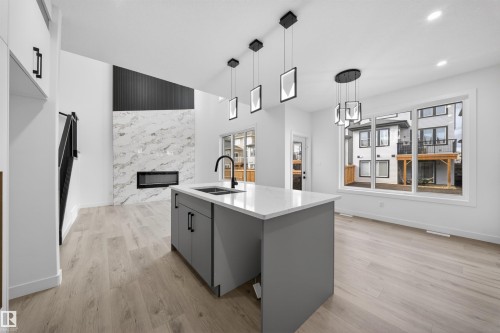 Open-concept space featuring a central island with a sink, light wood-finish flooring, a contemporary fireplace with a large-format stone surround, and multiple pendant light fixtures - 3906 41 Avenue, Beaumont, AB - Indoor Photo Showing Kitchen With Fireplace With Double Sink