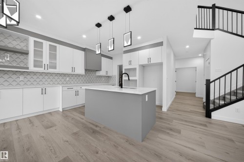 Open-concept kitchen featuring an island with a built-in sink, quartz countertops, white cabinetry with glass inserts, and wood-finish flooring - 3906 41 Avenue, Beaumont, AB - Indoor Photo Showing Kitchen With Upgraded Kitchen