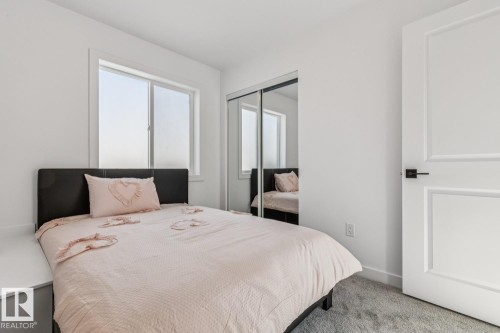 Bright bedroom featuring a window with white trim, built-in mirrored closet doors, a white paneled door with black hardware, plush gray carpeting, and white walls - 10726 107 Street, Edmonton, AB - Indoor Photo Showing Bedroom