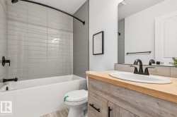 Modern bathroom featuring a white bathtub and toilet, light gray subway tile surround, matte black fixtures, and a vanity with a light wood-finish countertop and sink - 