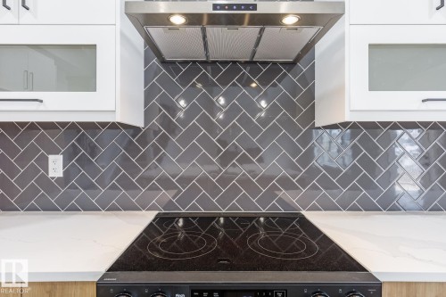 Modern kitchen featuring a herringbone pattern subway tile backsplash, white upper cabinetry with frosted glass inserts, and a stainless steel range hood - 917 Elderberry Landing, Edmonton, AB - Indoor
