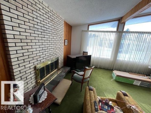 Living area featuring an exposed brick fireplace with a brass-toned firebox and hearth, green carpeting, and large windows with vertical blinds - 11003 144 Street, Edmonton, AB - Indoor With Fireplace