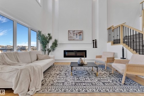 Living area featuring soaring ceilings, an integrated linear electric fireplace, large picture windows, a wood-finish staircase with black metal balusters, and light wood-finish flooring - 247 Linden Loop, Leduc, AB - Indoor Photo Showing Living Room With Fireplace