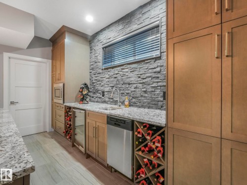 Wet bar featuring a textured stone accent wall, granite countertops, wood-finish cabinetry, built-in wine storage, and stainless steel appliances - 8927 120 Street, Edmonton, AB - Indoor Photo Showing Kitchen