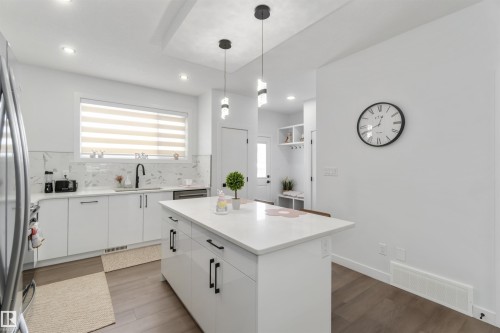 Bright kitchen featuring a central island with a solid surface countertop, white cabinetry with black hardware, a tile backsplash, and wood-finish flooring - 124 28 Street, Edmonton, AB 