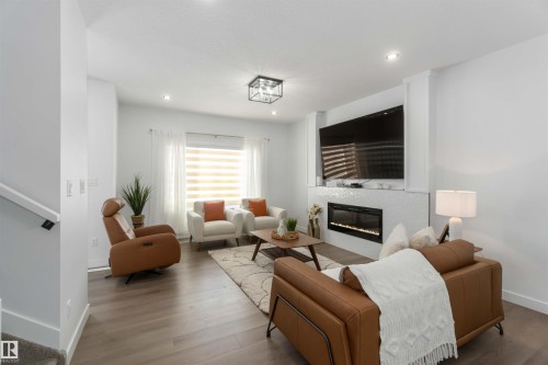 Living area featuring wood-finish flooring, a contemporary fireplace with white textured tile surround, recessed lighting, and a window with horizontal blinds and sheer drapes - 124 28 Street Sw, Edmonton, AB - Indoor Photo Showing Living Room With Fireplace
