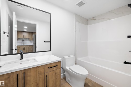 Bathroom featuring a wood-finish vanity with a white countertop and integrated sink, a large framed mirror, a tub/shower combination with a white surround and tile accent, and matte black fixtures - 954 Elderberry Landing, Edmonton, AB - Indoor Photo Showing Bathroom