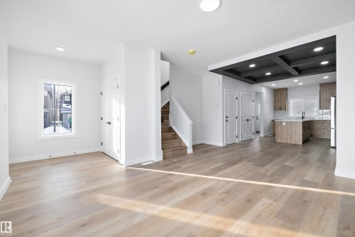 Open-concept main level featuring light wood-finish flooring, white trim, and a contemporary kitchen with wood-finish cabinetry, a central island, and a dark coffered ceiling - 954 Elderberry Landing, Edmonton, AB - Indoor Photo Showing Other Room
