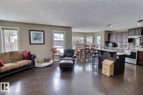 This open-concept living space features dark wood flooring, a kitchen with dark cabinetry and stainless steel appliances, and a dining area with ample natural light from surrounding windows - 1212 Westerra Crescent, Stony Plain, AB - Indoor