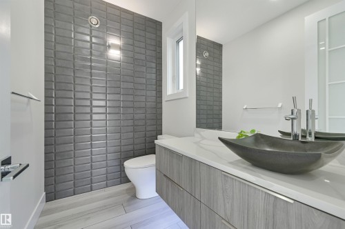 Contemporary bathroom featuring a textured gray accent wall, a floating wood-grain vanity with a white countertop, a modern vessel sink with chrome fixtures, a large wall-mounted mirror, and light-toned tile flooring - 9741 90 Avenue, Edmonton, AB - Indoor Photo Showing Bathroom