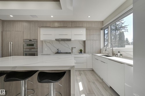 Modern kitchen featuring a large island with white quartz countertop, wood-finish flooring, integrated refrigerator, double wall ovens, and expansive window - 9741 90 Avenue, Edmonton, AB - Indoor Photo Showing Kitchen With Upgraded Kitchen