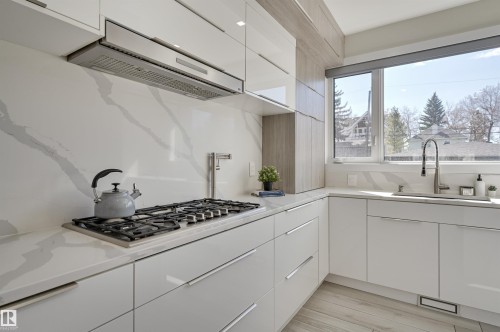 Gourmet kitchen featuring a built-in gas range, stainless steel range hood, stone countertops with matching backsplash, white flat-panel cabinetry, and a large window above the sink - 9741 90 Avenue, Edmonton, AB - Indoor Photo Showing Kitchen