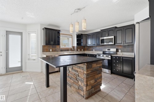 The property features a kitchen with dark cabinetry, stainless steel appliances, and a stone-clad island - 6009 36 Avenue, Beaumont, AB - Indoor Photo Showing Kitchen
