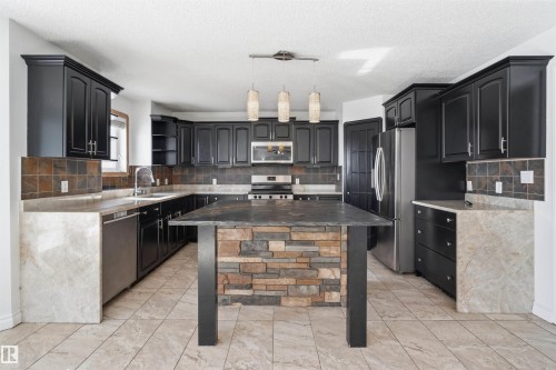 The kitchen features extensive dark cabinetry, an island with a stone base, stainless steel appliances, and a tiled backsplash - 6009 36 Avenue, Beaumont, AB - Indoor Photo Showing Kitchen
