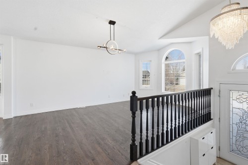 The property features dark wood flooring, a decorative black handrail, a contemporary chandelier, and large windows, including an arched window, providing natural light - 6009 36 Avenue, Beaumont, AB - Indoor Photo Showing Other Room