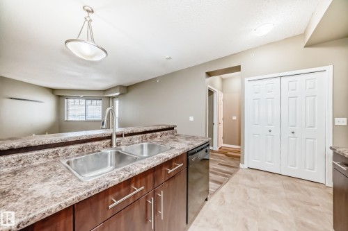 The kitchen features a double stainless steel sink with a high arc faucet, dark wood cabinetry with chrome handles, and a patterned countertop - 411 6925 199 Street Nw, Edmonton, AB - Indoor Photo Showing Kitchen With Double Sink