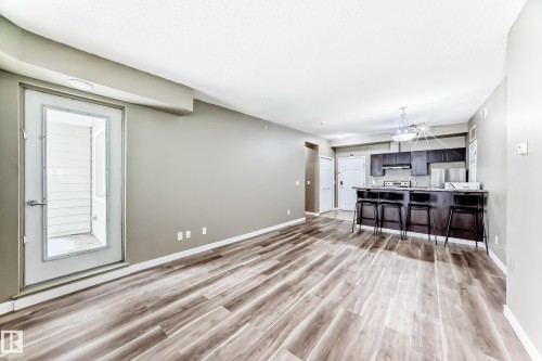Open concept living space featuring wood-look flooring, a kitchen island with seating, dark wood cabinetry, and a full-light door leading to an outdoor area - 411 6925 199 Street Nw, Edmonton, AB - Indoor