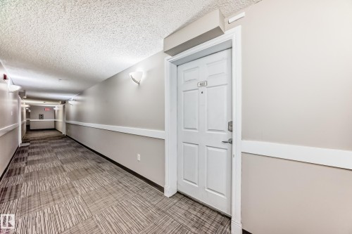 The property offers convenient access via a well-maintained corridor featuring patterned carpeting, neutral-toned walls, and wall-mounted lighting fixtures - 411 6925 199 Street Nw, Edmonton, AB - Indoor Photo Showing Other Room