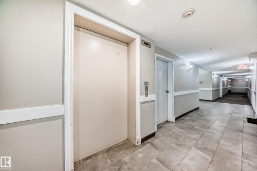 The property features an elevator with call buttons, a hallway with tiled flooring, and wall-mounted lighting fixtures - 411 6925 199 Street Nw, Edmonton, AB - Indoor Photo Showing Other Room
