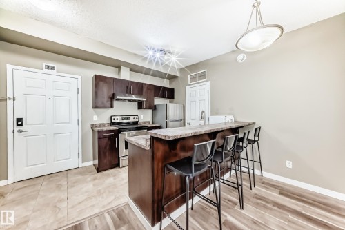 The property features a kitchen with dark wood cabinetry, stainless steel appliances, and a breakfast bar with a stone countertop - 411 6925 199 Street Nw, Edmonton, AB - Indoor Photo Showing Kitchen