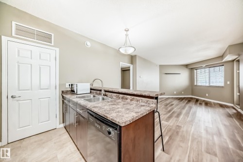 Open concept living area featuring a kitchen with a double basin sink, stainless steel dishwasher, and ample counter space, alongside a living room with wood-look flooring and a window with blinds - 411 6925 199 Street Nw, Edmonton, AB - Indoor Photo Showing Kitchen With Double Sink