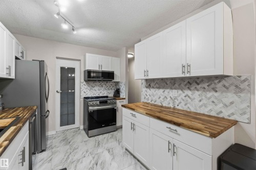 Kitchen featuring white shaker-style cabinetry, wood-finish countertops, stainless steel appliances, herringbone tile backsplash, and large format floor tiles - 5740 172 Street, Edmonton, AB - Indoor Photo Showing Kitchen