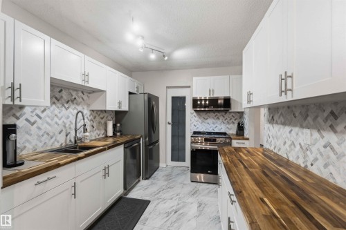 Modern kitchen featuring white shaker cabinetry, stainless steel appliances, wood-finish countertops, herringbone tile backsplash, and marble-look tile flooring - 5740 172 Street, Edmonton, AB - Indoor Photo Showing Kitchen With Double Sink