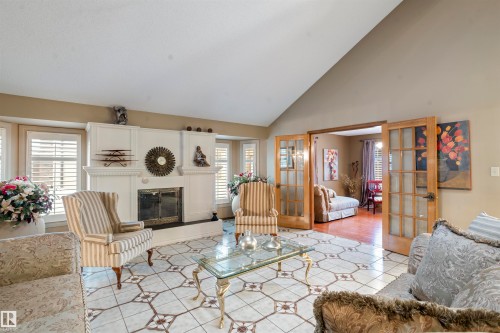 Vaulted ceiling living space featuring a white fireplace with an ornate mantel, patterned tile flooring, multiple windows with white plantation shutters, and light wood French doors with glass panels - 9831 158 Avenue, Edmonton, AB - Indoor Photo Showing Living Room With Fireplace