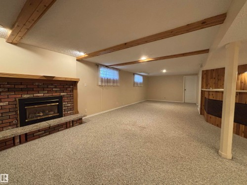 Spacious room featuring exposed wood ceiling beams, a brick fireplace with a wood mantle and stone hearth, plush carpet flooring, recessed lighting, and a wood-paneled feature wall - 11139 30 Avenue, Edmonton, AB - Indoor Photo Showing Basement With Fireplace