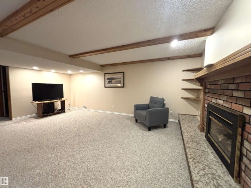 Spacious carpeted room featuring a brick fireplace with a wood mantel, exposed ceiling beams, recessed lighting, and built-in shelving - 11139 30 Avenue, Edmonton, AB - Indoor Photo Showing Basement