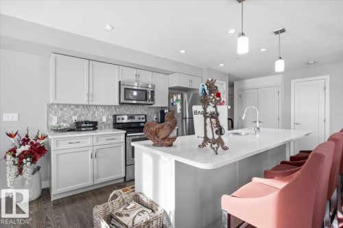 The kitchen features white shaker style cabinetry, stainless steel appliances, a tiled backsplash, and a large island with seating - 211 5029 Edgemont Boulevard, Edmonton, AB - Indoor Photo Showing Kitchen With Upgraded Kitchen