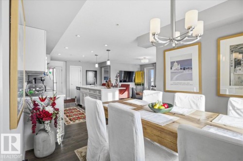 The dining area features a wooden table with white chairs, illuminated by a modern chandelier - 211 5029 Edgemont Boulevard, Edmonton, AB - Indoor Photo Showing Dining Room
