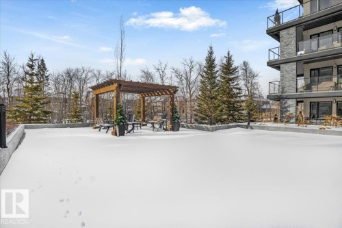 This outdoor space features a wooden pergola with seating, surrounded by evergreen trees and a building with balconies - 211 5029 Edgemont Boulevard, Edmonton, AB - Outdoor With Balcony