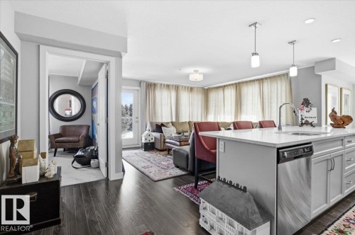 The property features hardwood floors, a kitchen island with a sink and pendant lighting, and a living area with large windows providing natural light - 211 5029 Edgemont Boulevard, Edmonton, AB - Indoor Photo Showing Kitchen With Upgraded Kitchen