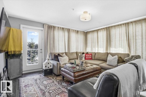 Living area featuring a full-light door with a view of snow-covered evergreens, dark flooring, and a ceiling light fixture - 211 5029 Edgemont Boulevard, Edmonton, AB - Indoor Photo Showing Living Room