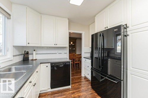 Bright kitchen featuring white cabinetry, dark countertops, a double basin stainless steel sink, black appliances, and wood-finish flooring - 1727 65 Street, Edmonton, AB - Indoor Photo Showing Kitchen With Double Sink