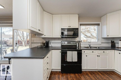 Kitchen featuring white cabinetry, dark countertops, wood-finish flooring, and black appliances - 1727 65 Street, Edmonton, AB - Indoor Photo Showing Kitchen With Double Sink