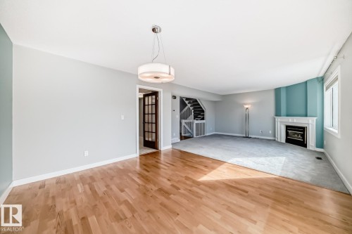 Spacious room featuring wood-finish flooring, a contemporary ceiling light fixture, a fireplace with a white mantel, and a staircase with white railings - 2709 124 Street, Edmonton, AB - Indoor Photo Showing Other Room With Fireplace