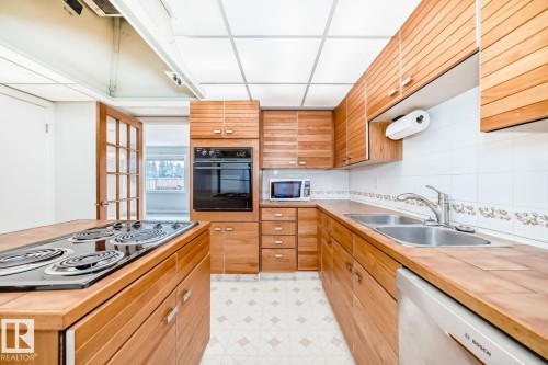 Kitchen featuring wood-finish cabinetry, integrated wall oven, dual basin stainless steel sink, and a built-in cooktop on a central island - 2709 124 Street, Edmonton, AB - Indoor Photo Showing Kitchen With Double Sink