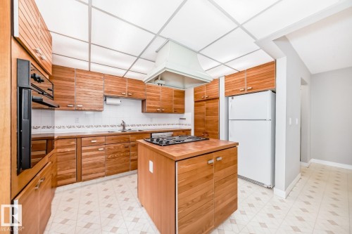 Kitchen featuring extensive wood cabinetry, a built-in oven, a center island with a cooktop, a white tile backsplash, and patterned vinyl flooring - 2709 124 Street, Edmonton, AB - Indoor Photo Showing Kitchen With Double Sink