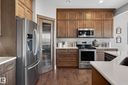 Modern kitchen featuring stainless steel appliances, wood-finish cabinetry, light-toned countertops, tile backsplash, and wood-finish flooring - 2086 Redtail Common, Edmonton, AB - Indoor Photo Showing Kitchen