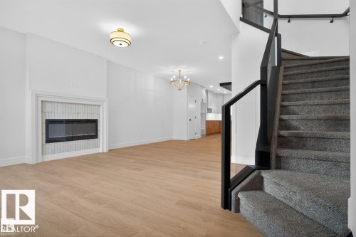 Open-concept living area with a contemporary electric fireplace featuring a textured tile surround, wood-finish flooring, a modern chandelier, and a carpeted staircase with a black handrail and glass panels - 55 Rosa Crescent, St. Albert, AB - Indoor With Fireplace