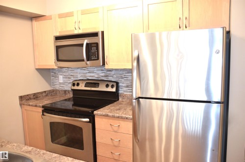 Kitchen with wood-finish cabinetry, stainless steel appliances, speckled countertops, and a mosaic tile backsplash - 103 920 156 Street Nw, Edmonton, AB - Indoor Photo Showing Kitchen